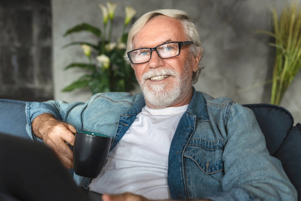 Portrait of smiling kind elderly 60s man sitting relaxing on the couch with cup of coffee. Happy pension - Fotografie, Obrázek