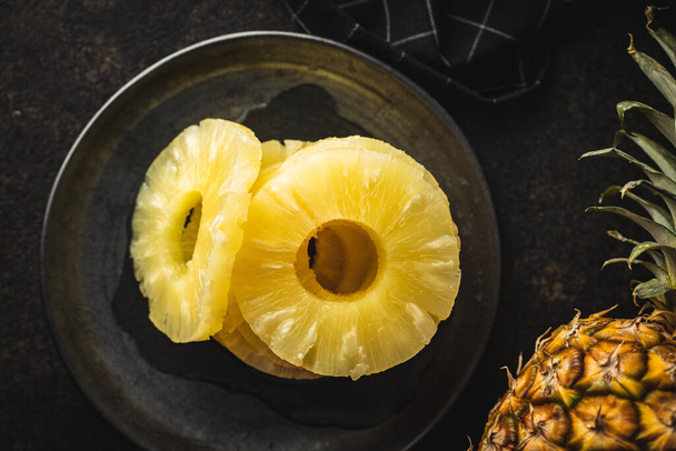 Canned sliced pineapple fruit on plate on black table. Top view. - 写真・画像