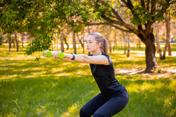 Young attractive woman dressed in sports uniform doing squat plank with dumbbells in hands. Sports activities in the park. Healthy lifestyle concept. - Fotó, kép