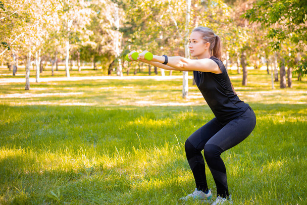Young attractive woman dressed in sports uniform doing squat plank with dumbbells in hands. Sports activities in the park. Healthy lifestyle concept. - Valokuva, kuva