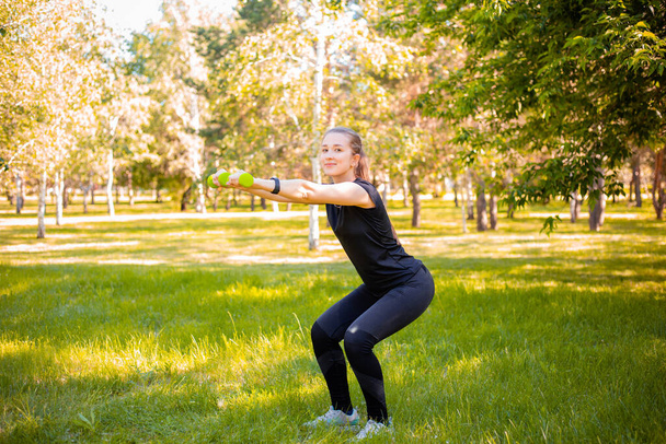 Young attractive woman dressed in sports uniform doing squat plank with dumbbells in hands. Sports activities in the park. Healthy lifestyle concept. - Foto, Imagen