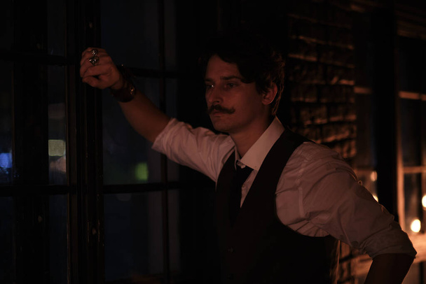 Şık giyinmiş bir adam pencerede durur ve mesafeye bakar. Düşünceli adam gece karanlık bir odada duruyor. - Fotoğraf, Görsel