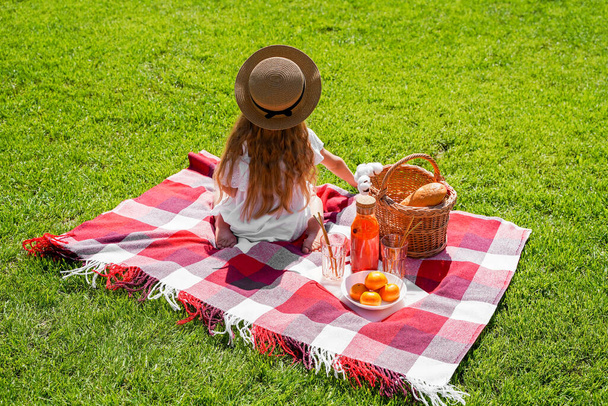 Дівчина з довгим волоссям у капелюсі сидить зі спиною, пікнік у парку на зеленій траві опівдні, шкільні канікули, відпустка та подорожі
. - Фото, зображення