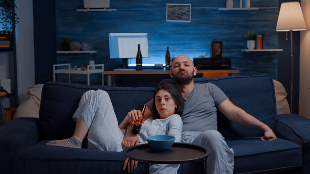 Расслабься, пара в пижаме сидит на диване и ест попкорн перед телевизором - Фото, изображение