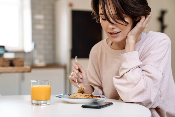 Tyytyväinen kaunis tyttö ottaa aamiaisen istuessaan pöydässä kotona - Valokuva, kuva