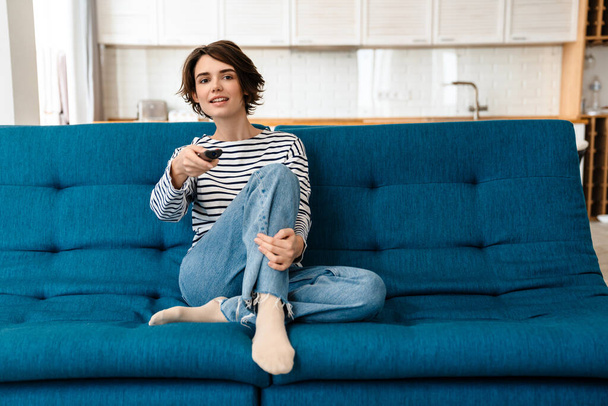 Happy beautiful woman smiling and using remote control while watching TV at home - Фото, изображение