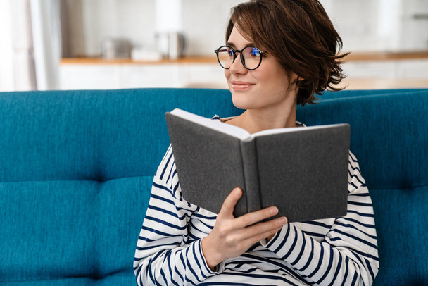 Happy beautiful woman in eyeglasses reading book while sitting on couch at home - Fotografie, Obrázek