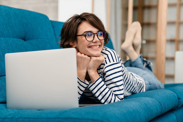 Happy beautiful woman in eyeglasses using laptop while lying on couch at home - Fotó, kép