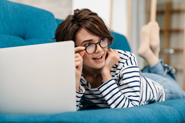 Happy beautiful woman in eyeglasses using laptop while lying on couch at home - Foto, Imagem