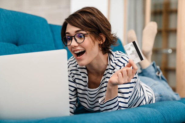 Excited beautiful woman in eyeglasses using credit card and laptop on couch at home - Foto, Bild