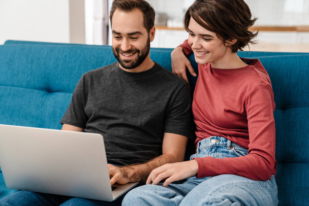 Happy young couple smiling and using laptop while sitting on couch at home - Fotoğraf, Görsel