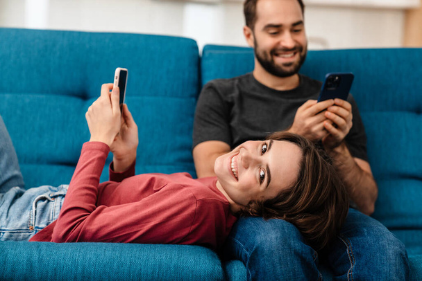 Happy young couple smiling and using mobile phones while resting on couch at home - Fotó, kép