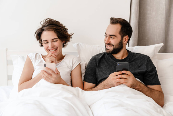 Happy young couple smiling and using mobile phones while lying in bed at home - Fotó, kép