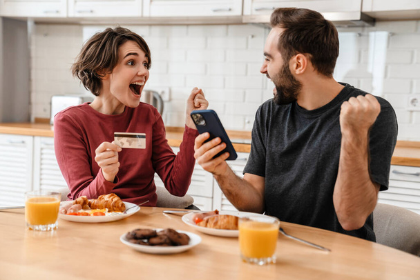 Excited couple making winner gesture using cellphone and credit card at home kitchen - Fotó, kép