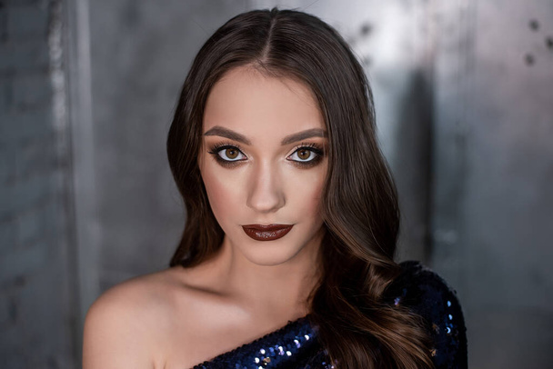Close-up portrait of beautiful girl with an evening festive professional make-up in blue, gold dress in sequins on metallic, gray shiny background. Grunge texture of interior. Clean skin, healthy face - Foto, imagen