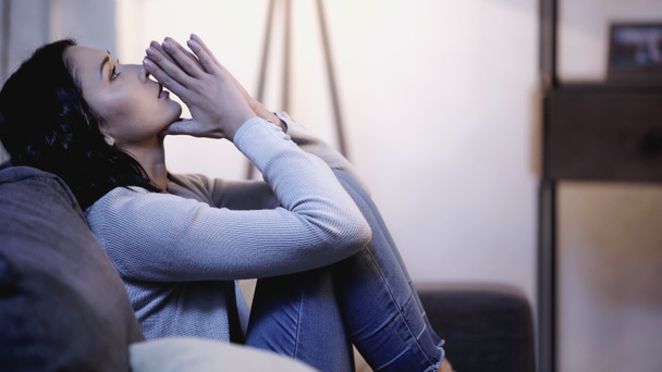 mulher deprimida em roupas casuais sentado no sofá com as mãos de oração perto da boca e olhando para cima em casa - Foto, Imagem