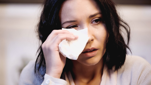 portrait of crying brunette woman wiping tears with white paper napkin at home - Photo, Image