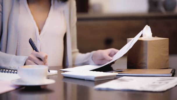 частковий вид жінки, що сидить на столі, тримає документи і пише з ручкою в блокноті вдома
 - Фото, зображення