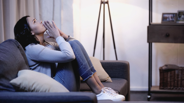 Depressive Frau in lässiger Kleidung sitzt mit betenden Händen in Mundnähe auf dem Sofa zu Hause - Foto, Bild