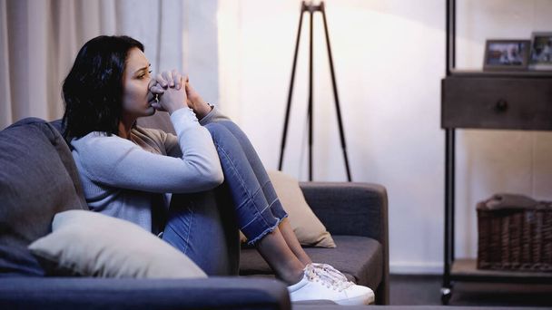 mujer deprimida en ropa casual sentada en el sofá y sosteniendo las manos apretadas cerca de la boca en casa - Foto, imagen