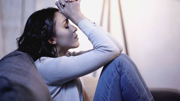 depressed woman in casual clothes sitting on sofa with closed eyes and holding clenched hands near head at home - Foto, Imagem