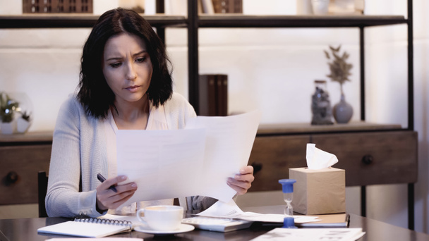 worried woman sitting on table and reading documents with pen in hand at home - Photo, Image