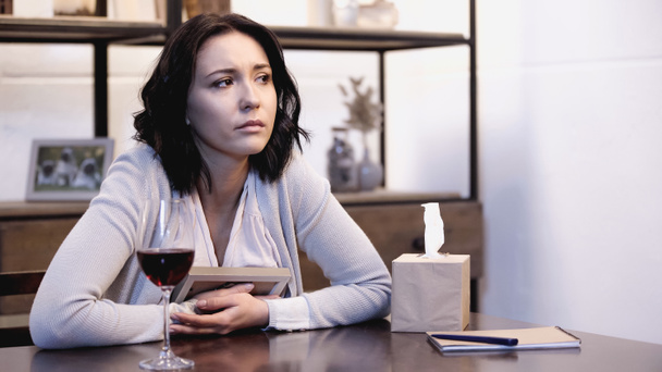 Aufgebrachte Frau sitzt auf Tisch und weint mit Fotorahmen in der Hand zu Hause - Foto, Bild