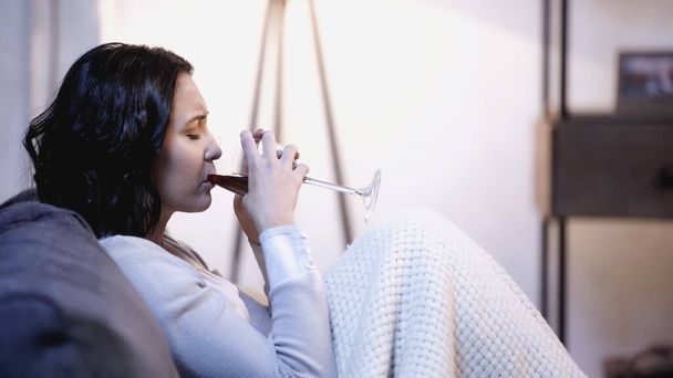 расстроенная женщина, покрытая одеялом, сидит на диване и пьет красное вино из бокала дома - Фото, изображение