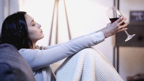 расстроенная женщина, покрытая одеялом, сидит на диване с бокалом вина в протянутых руках дома - Фото, изображение