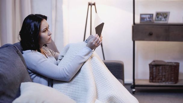 upset woman covered in blanket sitting on sofa and looking at envelope in hand at home - Photo, Image