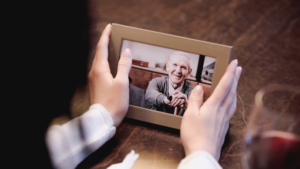 částečný pohled na ženské ruce držící fotorámeček s obrázkem staršího muže doma - Fotografie, Obrázek