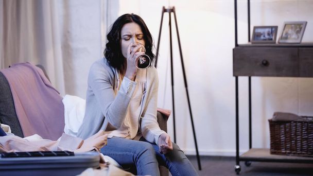 depressed woman sitting on sofa near male clothes and drinking red wine from glass at home - Photo, Image