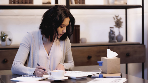 серьезная женщина сидит на столе и читает документы с ноутбуком и ручкой в руке дома - Фото, изображение