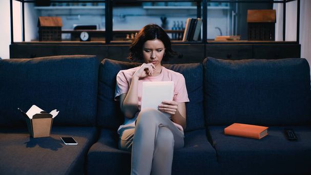 donna bruna che utilizza tablet digitale mentre è seduta sul divano vicino a smartphone, libro e scatola di cartone con cibo  - Foto, immagini