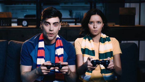 KYIV, UKRAINE - JUNE 09, 2021: man and woman in striped scarfs holding joysticks and playing video game  - Фото, изображение