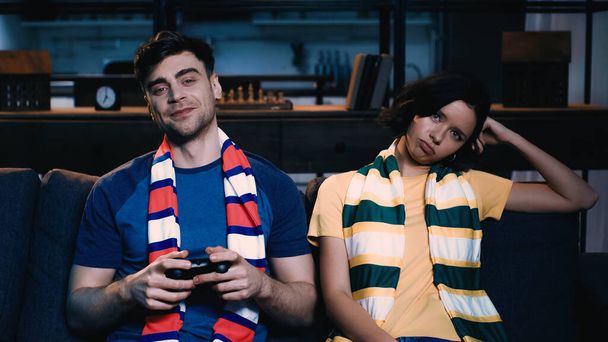 KYIV, UKRAINE - JUNE 09, 2021: cheerful man in striped scarf holding joystick and playing video game near displeased girlfriend  - Foto, imagen