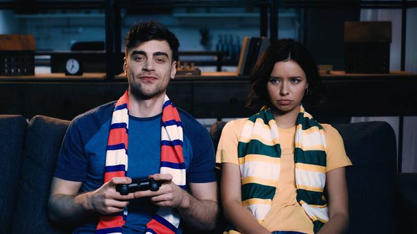 KYIV, UKRAINE - JUNE 09, 2021: cheerful man in striped scarf holding joystick and playing video game near offended girlfriend  - Foto, Imagen