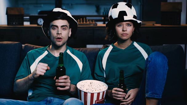 ファンの帽子のサッカーファンはビールのボトルを保持し、ソファの上でチャンピオンシップを見ます拳を食いしばり  - 写真・画像