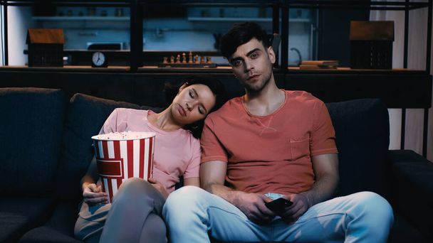 tired woman holding popcorn bucket and sleeping on shoulder of boyfriend in living room  - Foto, afbeelding