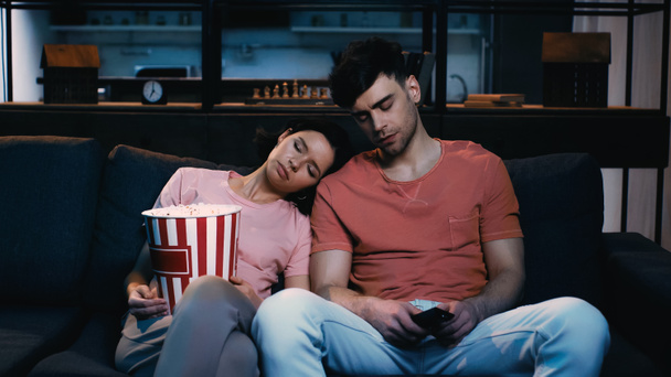 exhausted woman with popcorn bucket sleeping on shoulder of tired boyfriend in living room  - Фото, изображение