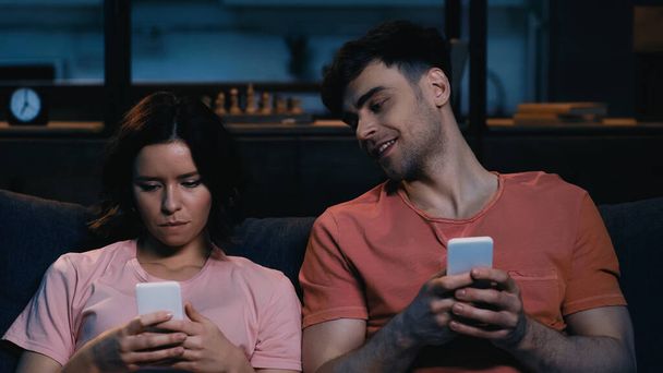 szczęśliwy mężczyzna patrząc na kobieta smsy na telefon komórkowy w nowoczesnym salonie  - Zdjęcie, obraz