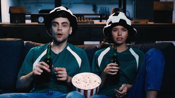 aficionados al fútbol tensos en sombreros de abanico sosteniendo botellas de cerveza y viendo el campeonato cerca de palomitas de maíz cubo  - Foto, Imagen