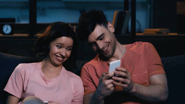 cheerful man and woman looking at smartphone and smiling in modern living room  - Фото, зображення