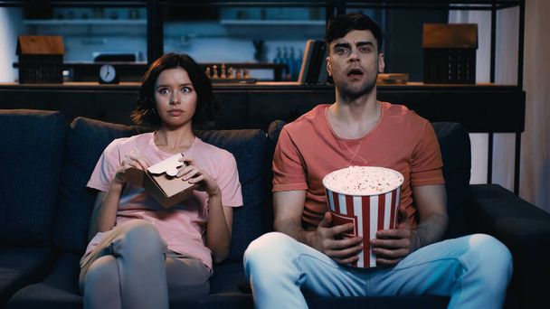 překvapený muž sleduje film a drží popcorn kbelík v blízkosti ženy sedí s lepenkovou krabicí  - Fotografie, Obrázek