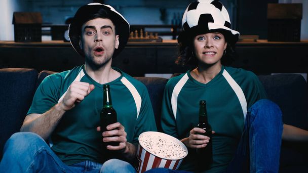 worried couple in fan hats watching championship and holding bottles of beer near popcorn bucket in living room clenched fist - Fotó, kép