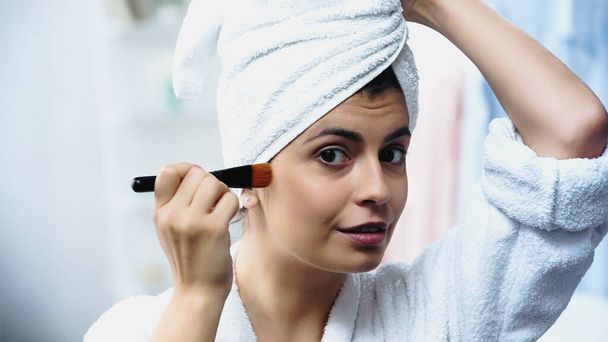 pretty young woman with head wrapped in towel applying foundation on face with cosmetic brush in bedroom - Photo, Image