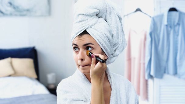 mujer joven con la cabeza envuelta en toalla aplicando la base de la cara bajo los ojos con cepillo cosmético en el dormitorio - Foto, imagen