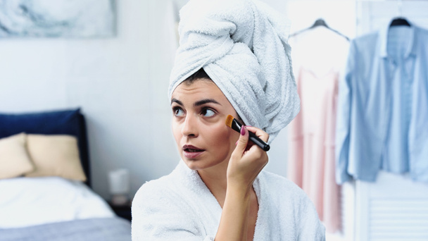 fiatal nő fürdőköntösben fej csomagolva törölköző alkalmazása alapítvány az arcon kozmetikai kefe a hálószobában - Fotó, kép