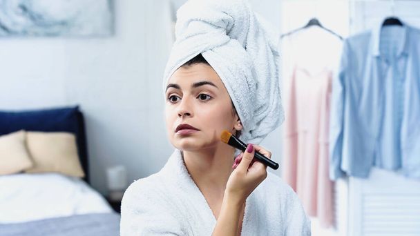 fiatal nő fürdőköntösben fej csomagolva törölköző alkalmazása alapítvány az arcon kozmetikai kefe a hálószobában - Fotó, kép