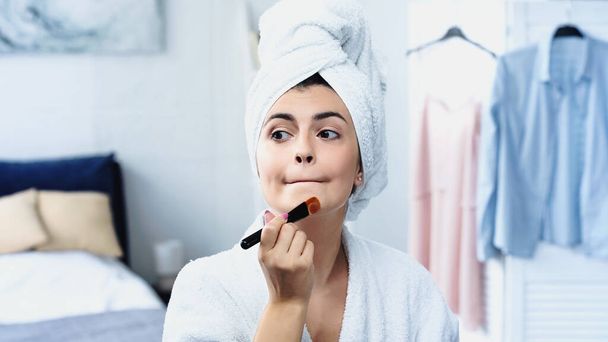 young woman in bathrobe with head wrapped in towel grimacing and applying face foundation in bedroom - Photo, Image